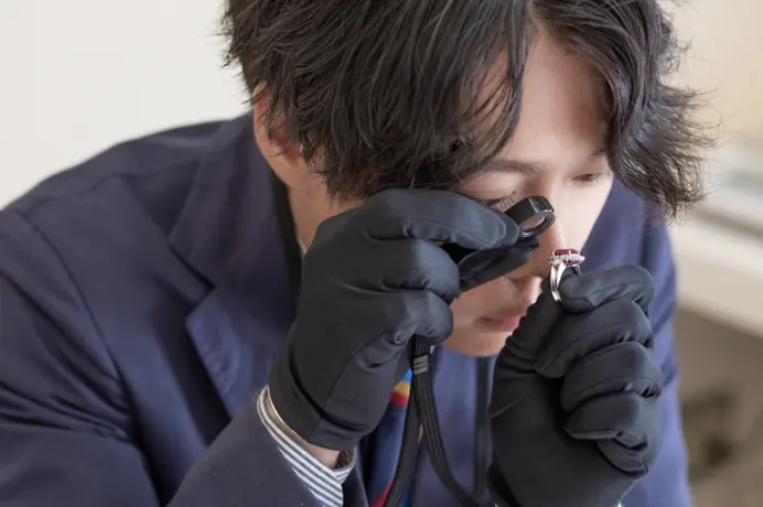 黒い手袋をはめ、ルーペを使って指輪の赤い宝石を慎重に鑑定する男性の姿。
