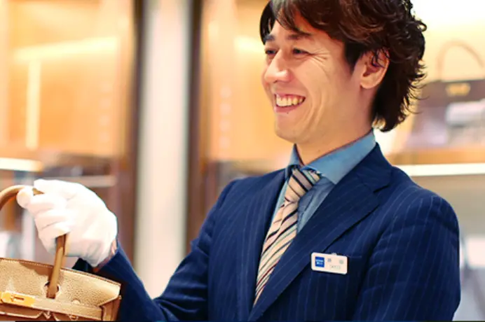 白い手袋をはめて高級ハンドバッグを持ち、笑顔を見せるスーツ姿の男性店員。