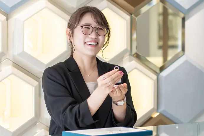 指輪を手に笑顔で接客する、眼鏡をかけた女性スタッフの姿。
